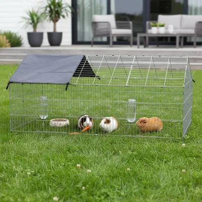 A wire cage for small animals is set up on a grassy lawn in front of a modern house with outdoor furniture.
