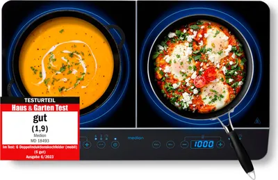 A black induction cooktop with two cooking zones, one with a pot of orange soup and the other with a pan of food with eggs and vegetables. A red and white test review badge is in the bottom left corner.