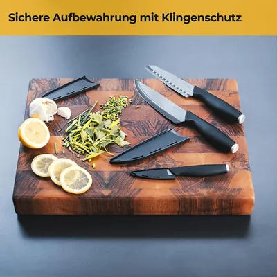 A set of three kitchen knives with black handles and protective sheaths are arranged on a wooden cutting board with chopped herbs, lemon slices, and a garlic bulb.