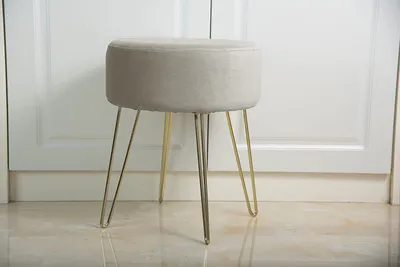 A beige upholstered stool with gold metal legs sits on a marble floor in front of a white door.