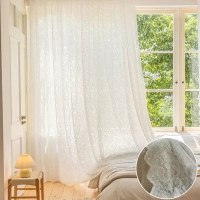 A bedroom scene with a sheer white curtain hanging in front of a window, with a bed and a small side table with a lamp visible.