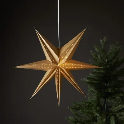 A gold, 7-pointed star lantern with a perforated pattern hangs in front of a dark background and a blurred Christmas tree.