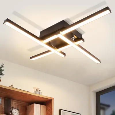 A modern LED ceiling light fixture with a geometric design, mounted on a white ceiling. The light is illuminated, casting shadows on the ceiling. Below the light, a wooden shelf with books and a clock is visible.