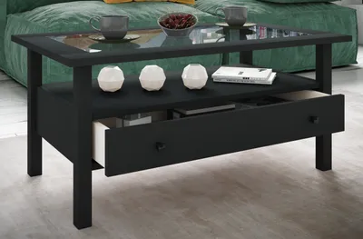 A black coffee table with a glass top and a drawer is shown in a living room setting, with a green sofa in the background.