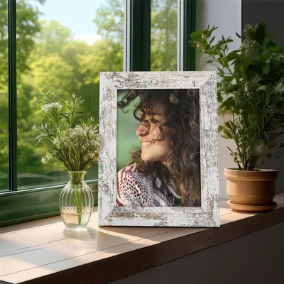 A framed photo of a woman sits on a windowsill next to a vase of flowers and a potted plant, with greenery visible through the window.