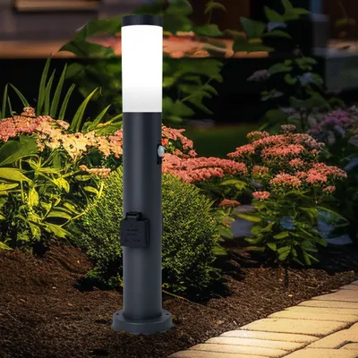A dark gray bollard light with a white top is illuminated in a garden setting at dusk, surrounded by plants and a paved walkway.