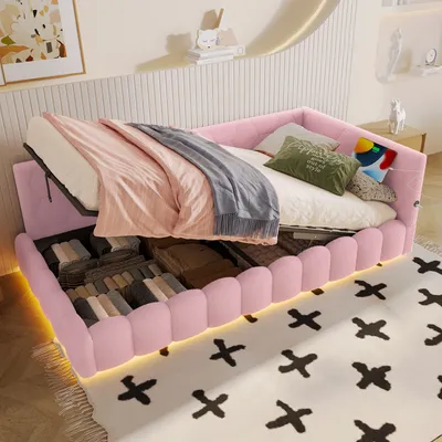 A pink upholstered bed with storage compartments open, revealing folded linens and blankets. The bed is made up with bedding and pillows.