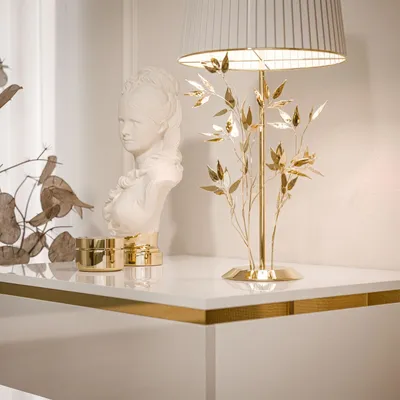 A white nightstand with a gold trim and a gold lamp with a white shade and a white bust on top.