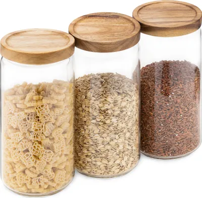 Three glass storage jars with wooden lids, filled with pasta, oats, and rice, are arranged side-by-side on a white background.