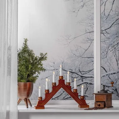 A wooden candle holder with seven electric candles is placed on a windowsill next to a small Christmas tree and a coffee grinder.
