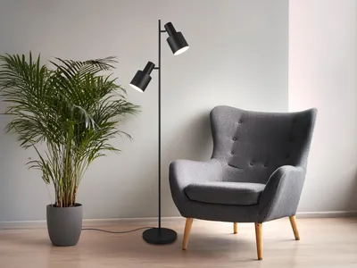 A black floor lamp with two adjustable spotlights stands next to a gray armchair and a large potted plant in a modern living room.