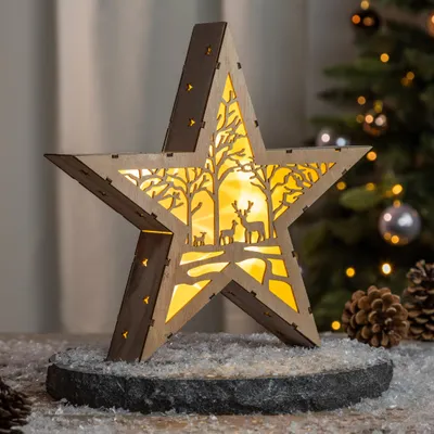 A wooden star-shaped LED light with a forest scene inside, placed on a stone base with faux snow.