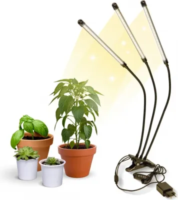 A plant grow light with three adjustable LED heads is positioned next to several potted plants on a white background.