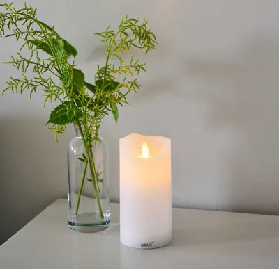 A white LED candle sits on a white surface next to a glass vase with green foliage.