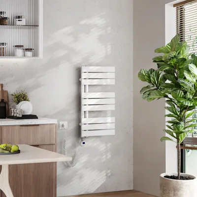 A modern kitchen or bathroom setting with a white electric towel warmer mounted on a light gray wall, next to a large potted plant and a wooden cabinet.