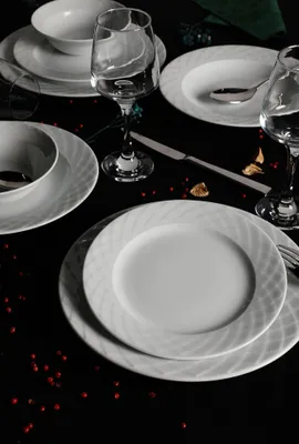 A formal table setting with white porcelain plates, bowls, and wine glasses on a black tablecloth, decorated with red peppercorns.