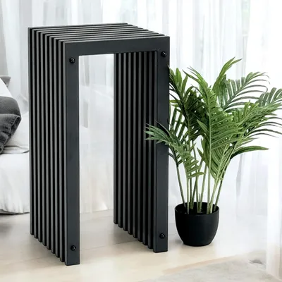 A black metal slatted stool is placed next to a potted plant in a room with sheer curtains in the background.