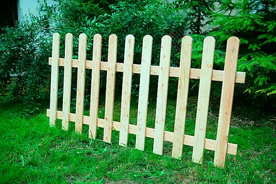 A wooden picket fence section is placed on a grassy lawn in front of green bushes and trees.