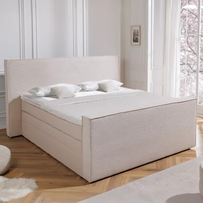 A beige boxspring bed with a headboard and mattress is shown in a well-lit room with wooden flooring and sheer curtains.