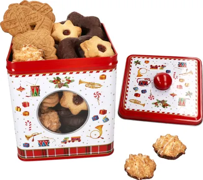 A vintage-style tin cookie box filled with various Christmas cookies, with the lid placed next to it. Two coconut macaroons are also shown in the foreground.