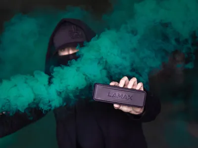 A person in a black hoodie and cap holds a dark rectangular object with the word "LAMAX" on it, surrounded by green smoke.