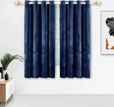 A pair of navy blue velvet curtains hangs on a curtain rod in front of a window. A small white cabinet with a wooden top is visible to the left, with a plant and a vase on it. A framed artwork is on the wall to the right.