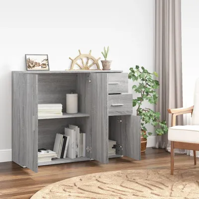A grey sideboard with open doors and drawers, displaying books and other items inside, set in a room with a plant and curtains.
