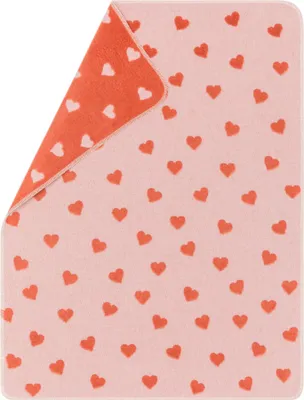 A pink baby blanket with small coral hearts scattered across it. The blanket is folded diagonally, showing a coral-colored underside with white hearts.