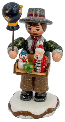 Ein handgefertigter Holzfigur zeigt einen Spielwarenhändler mit einem Tablett voller bunter Spielzeuge in den Händen und einem Vogel auf einem Stock während er auf einem schneebedeckten Sockel steht