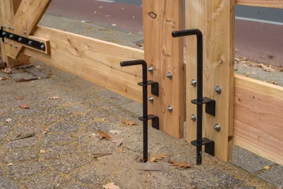 Close-up of two black metal gate latches attached to wooden posts, with a paved ground surface and fallen leaves in the foreground.
