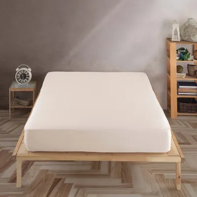 A bed with a beige fitted sheet on a wooden frame, next to a nightstand with an alarm clock and a wooden shelf unit with decorative items.