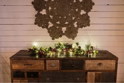 A rustic wooden dresser with a festive garland and lights draped across the top, set against a white wooden plank wall with a decorative jute rug hanging above.