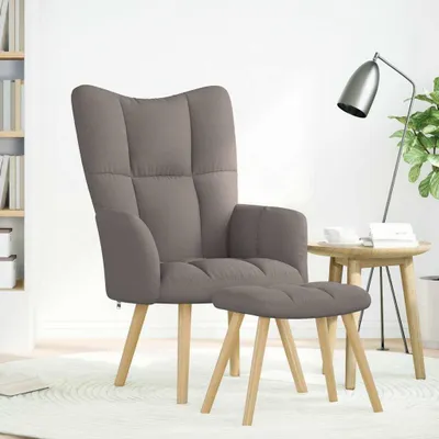A taupe fabric relaxsessel with a matching ottoman, placed on a white rug in a room with a bookshelf and a side table with a lamp and plant.