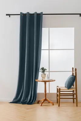 A blue velvet curtain hangs on a black rod next to a window. A small wooden table with a plant and a chair with a blue cushion are placed in front of the window.