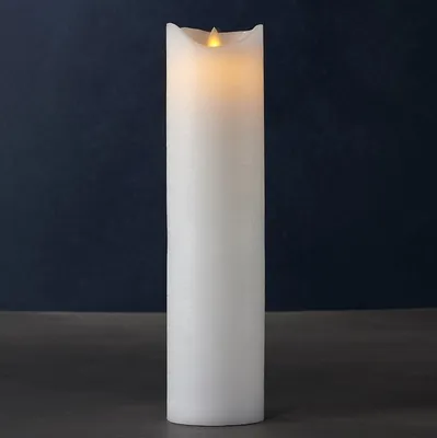 A tall, white LED candle with a flickering yellow flame sits on a dark surface against a dark blue background.