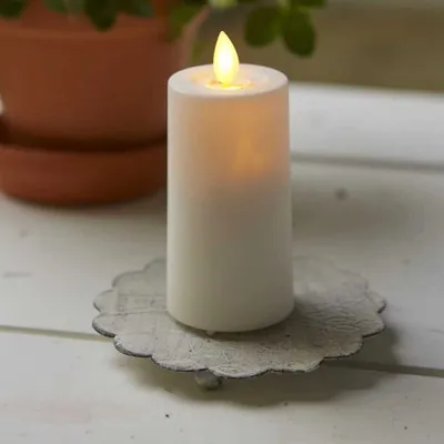 A white LED candle with a flickering yellow flame sits on a decorative metal candle holder. In the background, a terracotta pot with green plants is visible.