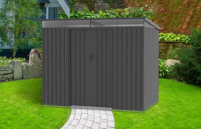 A dark gray metal shed with a skylight roof is situated in a grassy backyard with a stone pathway leading to its door. A brick wall and greenery form the background.