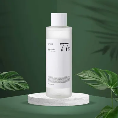 A bottle of Anua Heartleaf 77% Soothing Toner is displayed on a white circular stand, with lush green leaves and a dark green background.