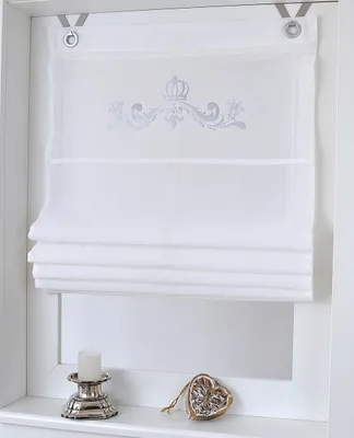 A white Roman blind with a silver crown motif is displayed in a window frame, with a candle holder and a decorative heart on the windowsill below.