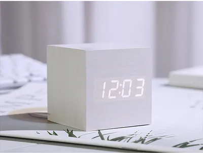 A white cube-shaped digital alarm clock with a bright LED display showing the time is placed on a desk with some papers and a keyboard in the background.