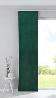 A green velvet blackout panel curtain hanging in front of a window with a view of trees and sky. A small white side table with a vase and decorative branches is to the left.