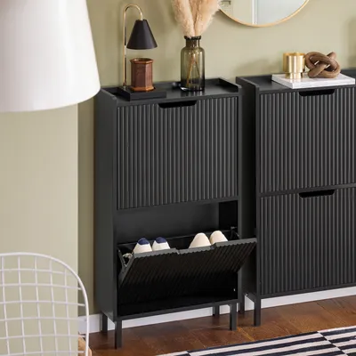 A black shoe cabinet with two flip-down drawers is shown in a room with a white chair and a lamp.