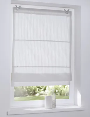 A white Roman blind with vertical stripes is shown hanging in a window. The blind is partially lowered, revealing a garden scene outside. A white cylindrical vase and a clear bottle are on the windowsill.