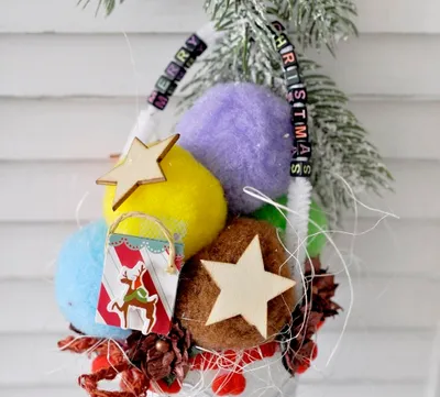A close-up of a Christmas decoration with colorful pom-poms, wooden stars, and a small gift bag.