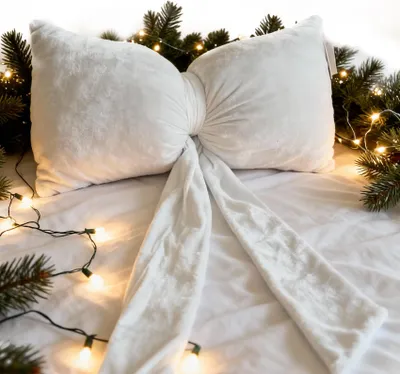 A white velvet bow-shaped pillow is displayed on a white surface, surrounded by pine branches and fairy lights, suggesting a festive or decorative setting.