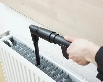 A hand is using a vacuum cleaner attachment to clean the fins of a white radiator.