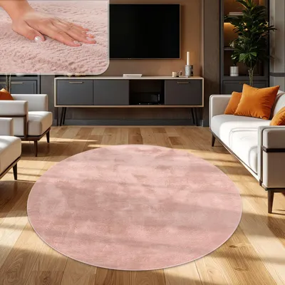 A round, pink rug is placed on a wooden floor in a modern living room. Furniture includes a sofa with orange cushions, armchairs, and a media console with a TV.