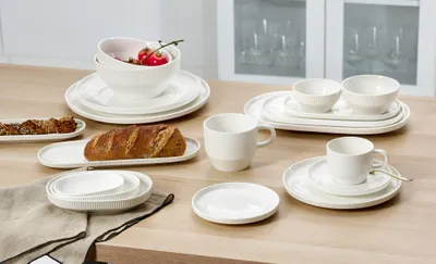 A collection of white ceramic tableware, including bowls, plates, and cups, arranged on a wooden table. Some bread and food items are also present.