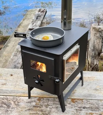A portable wood-burning stove is set up outdoors on a wooden deck, with a lake in the background. An egg is frying in a pan on top of the stove, and flames are visible inside the stove.