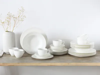 A collection of white porcelain dinnerware, including plates, bowls, and cups, arranged on a wooden shelf against a white background.